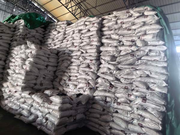 Towering stack of bagged industrial caustic soda stored under protective green tarpaulin in a warehouse