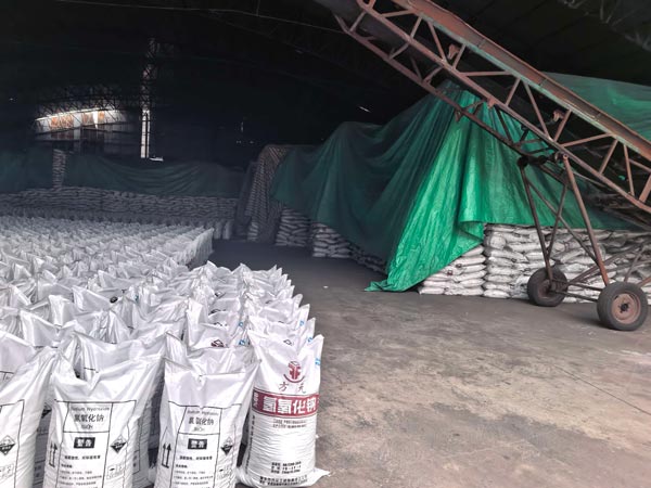 Mobile inclined conveyor placed beside stacked pure caustic soda bags under protective tarpaulin in a warehouse