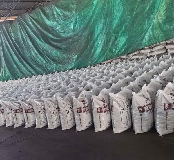 Massively stacked bulk caustic soda bags under protective factory tarpaulin ready for global export logistics.