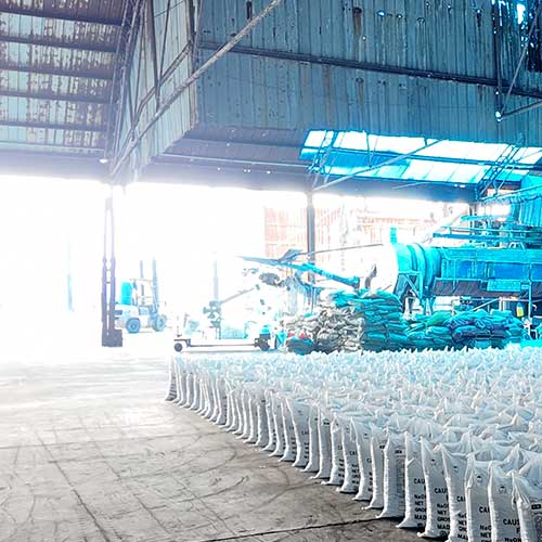 Foreground rows of 25kg caustic soda bags organized inside a chemical processing factory run by sodium hydroxide bulk suppliers.