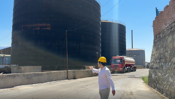 Plant auditor wearing hard hat gesturing toward outdoor bulk liquid storage tanks in soda caustic flake facility.